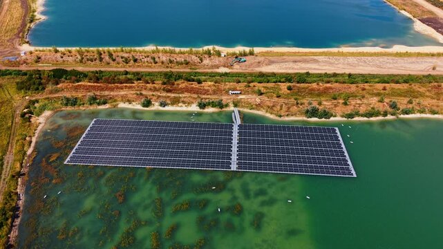 Floating photovoltaic array between two quarry ponds. Dark panel rows cover part of green water, with a dirt embankment separating the installation from another pond.