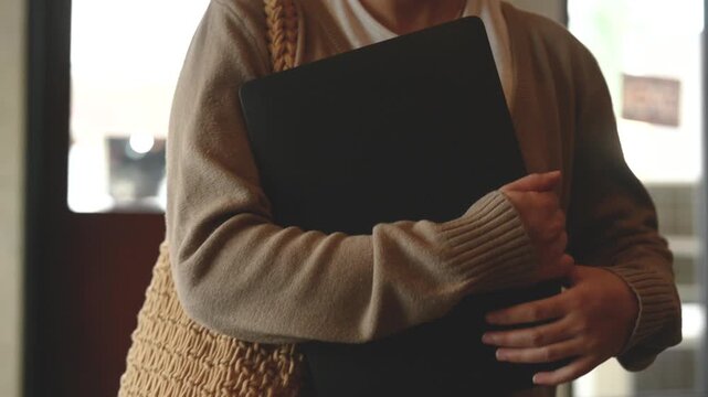 A woman is carrying books and a closed laptop against her torso near a cafe doorway. The woman walks past brickwork and tables with a woven shoulder bag at her side in soft daylight.