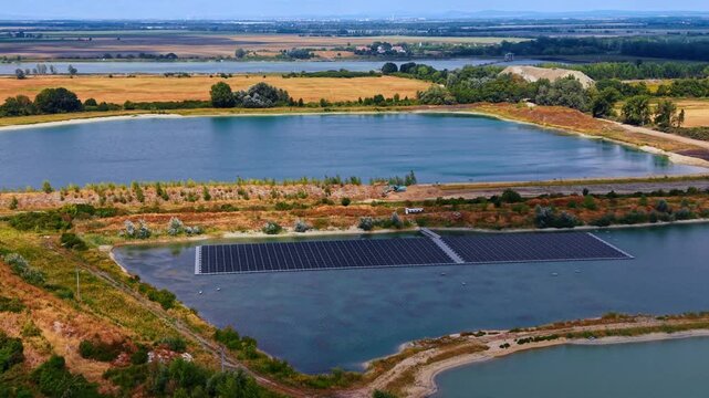 Floating solar farm across a pond in an agricultural landscape. Panel rows form a long rectangle on water, with embankments, a second lake and fields extending behind.