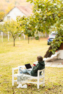 A woman sitting on a white wooden bench under an apple tree with a laptop and a red mug during autumn in a rural garden