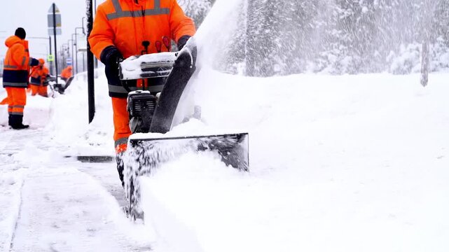 Snow clears swiftly with mechanical force. Workers in bright orange gear move steadily through falling flakes. Snowblowers push snow sideways, revealing cleared paths