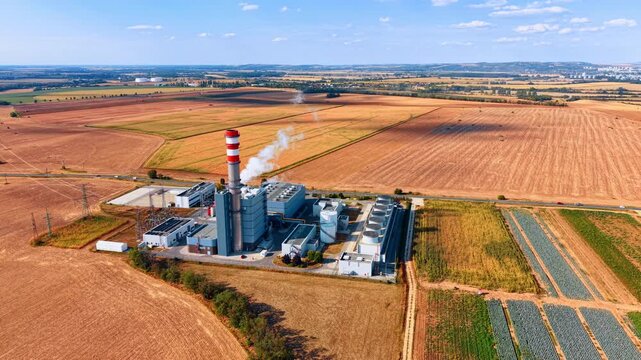 Power station within a patchwork of agricultural fields. A compact industrial complex emits white vapor beside a striped stack, with access roads and farmland around it.