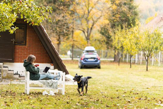 A woman sitting on a white wooden bench with a laptop and a red cup while a black dog lies nearby in a yard during autumn