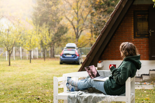 A person sitting outdoors on a wooden bench with a laptop during autumn in front of a rural house and a car.