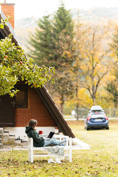A person sitting outdoors on a wooden bench with a laptop during autumn in front of a rural house and a car.