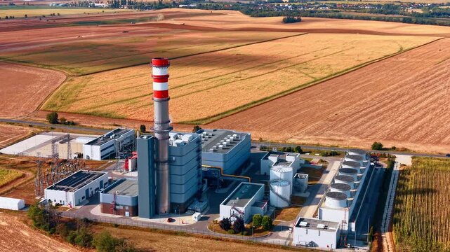 Power generation facility bordered by agricultural fields. A red and white chimney stands above gray buildings, cooling fans and tanks beside rows of electrical equipment.