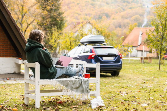 A person sitting outdoors on a wooden bench with a laptop during autumn in front of a rural house and a car.