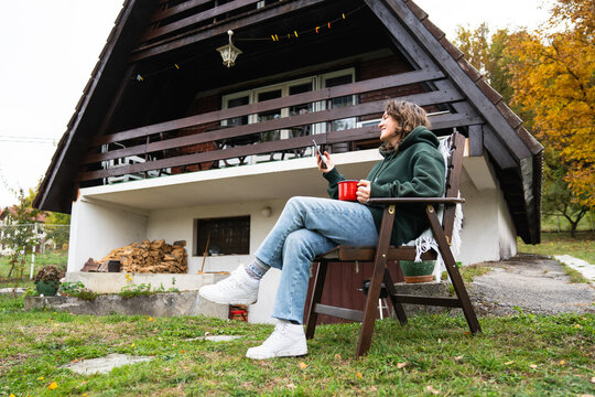 A woman sitting in a wooden chair outside an A-frame house holding a smartphone and a red mug during autumn