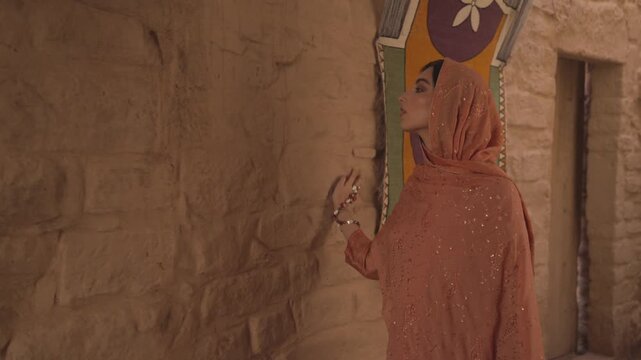 Cinematic medium shot of an elegant woman in traditional modest dress touching historic mud brick walls in AlUla Old Town