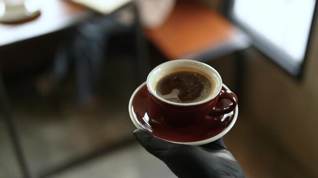 A coffee cup filled with a dark hot drink sits on a matching saucer at a cafe table. The coffee cup and saucer fill the close up, while the wooden tabletop and background fall softly out of focus.