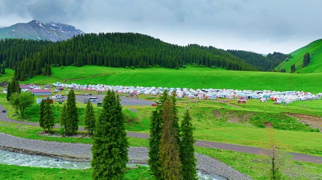 Scenic view of Tangbula Grassland in Ili, Xinjiang, featuring traditional yurts nestled between lush pine forests and alpine mountains under a cloudy sky.