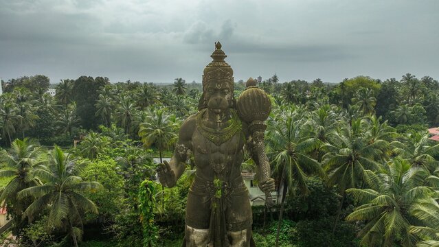 Aerial view of the large stone Hanuman statue holding a mace, surrounded by lush tropical palm trees under an overcast sky in Udupi, Karnataka, India.