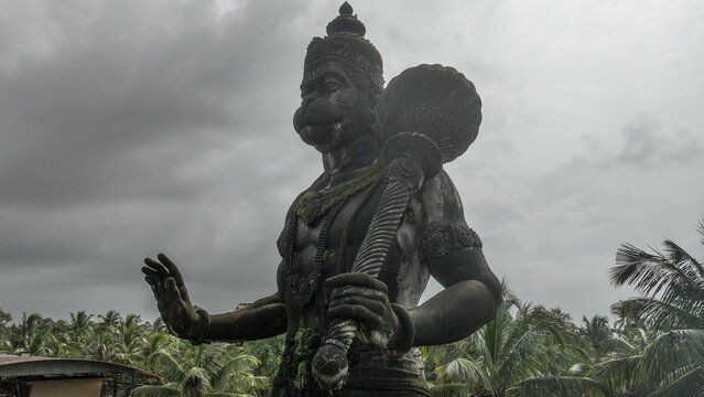 Aerial view of the stone statue of Lord Hanuman holding a mace surrounded by lush palm trees under an overcast sky in Udupi, Karnataka, India.