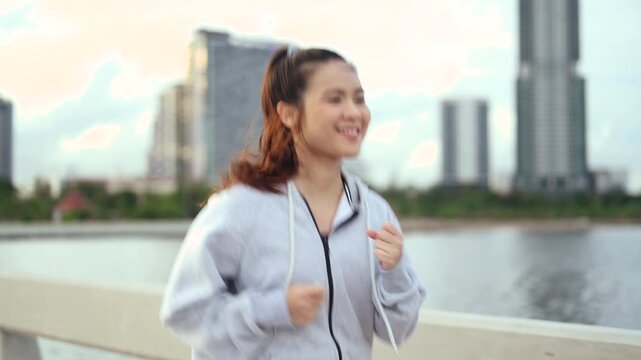 A woman is jogging along a waterfront path in sportswear, with bent arms and a steady running stride. The woman passes a railing while water and distant buildings stretch behind the pavement.