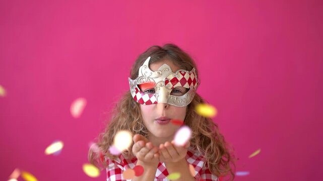 A happy child blows confetti from a handful. Beautiful little girl in a carnival mask on a pink background. Slow motion