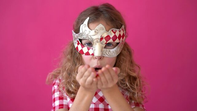 A happy child blows confetti from a handful. Beautiful little girl in a carnival mask on a pink background. Slow motion