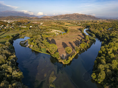 Aerial view of the Drin river winding through lush greenery and agricultural fields with mountains in the background in Shkoder, Shkoder County, Albania.