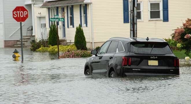 A black SUV sits partially submerged in high floodwater on a residential street, framed by a stop sign, fire hydrant and houses; standing water convey danger, disruption and storm damage.