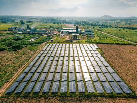 Aerial view of a solar farm with rows of photovoltaic panels, industrial buildings in the background, and surrounding rural landscape in Ogun State, Nigeria.