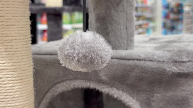 Fluffy grey pompom toy swinging on a rope attached to a faux fur cat tree house. Pet supplies retail store display, cozy animal entertainment accessory details.