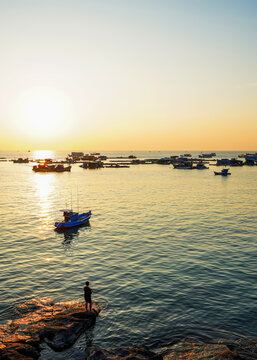 Picturesque sunset view of sea featuring fishing vessels and floating fishing villages off Phu Quoc Island, Vietnam. Silhouette of person with fishing rod standing on rocks by water's edge at dawn.