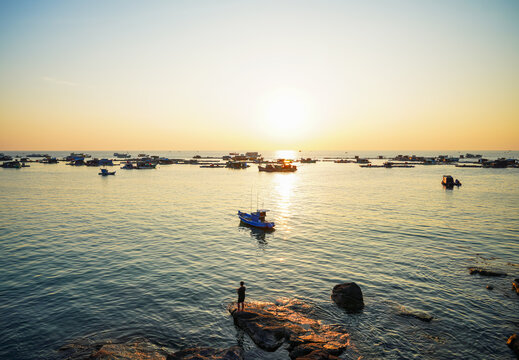 Picturesque sunset view of sea featuring fishing vessels and floating fishing villages off Phu Quoc Island, Vietnam. Silhouette of person with fishing rod standing on rocks by water's