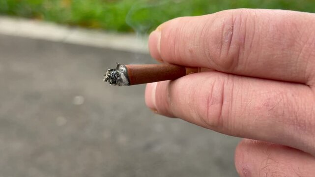 Close-up of a man hand holding a burning brown cigarette butt with ash outdoors. Unhealthy smoking habit background, nicotine addiction concept, toxic cigarette smoke details.
