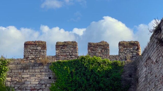 Timelapse of white clouds moving quickly across blue sky behind ancient fortress crenellations in Istanbul, Turkey. Ivy-covered stone wall forming historic Ottoman landmark - time lapse