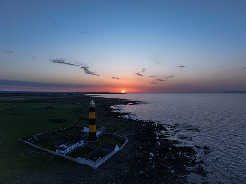 Aerial view of St John's Point Lighthouse standing on the rocky coastline during a vibrant sunset with green fields and the sea in Newcastle, Northern Ireland, United Kingdom.