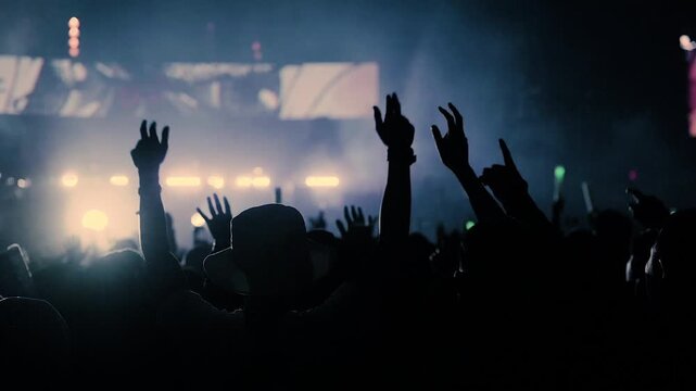 A dark stage shows bright screens beyond audience silhouettes in a low light scene. The dark stage is hidden by the crowd, while rectangular panels and small lights punctuate the black background.