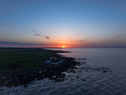 Aerial view of St John's Point Lighthouse at sunset with rocky coastline, green fields, and calm sea in Newcastle, Northern Ireland, United Kingdom.