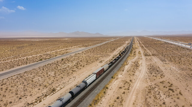 Aerial view of a long freight train traveling through the vast arid desert landscape with a parallel highway under a clear blue sky in the United States.