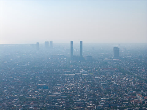 Aerial view of the dense urban sprawl, featuring residential rooftops, prominent high-rise towers, and hazy atmospheric conditions in Surabaya, East Java, Indonesia.