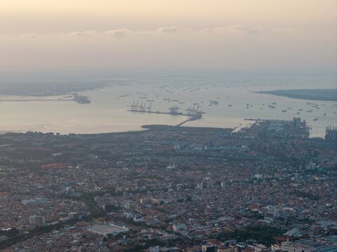 Aerial view of the port of Surabaya with industrial cranes and numerous ships in the bay, overlooking the dense urban sprawl of the city in Surabaya, East Java, Indonesia.