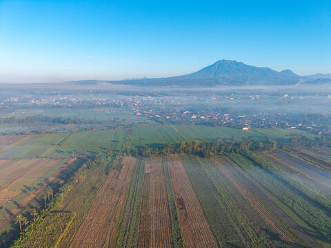 Aerial view of agricultural fields and mist-covered town with a prominent mountain in the background in Kepanjen, East Java, Indonesia.