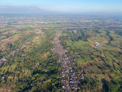 Aerial view of the town of Malang surrounded by lush green agricultural fields and rolling hills under a clear sky, Malang, East Java, Indonesia.