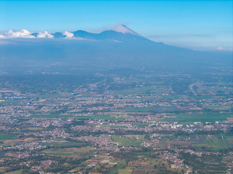 Aerial view of Mount Semeru rising above the city, surrounded by lush green fields, agricultural land, and urban sprawl in Malang, East Java, Indonesia.