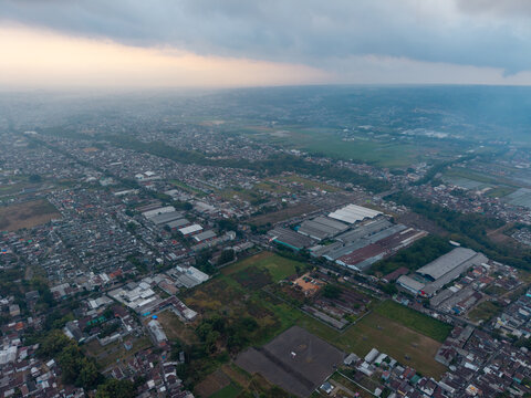 Aerial view of the urban landscape featuring dense residential areas, industrial warehouses, and surrounding green fields in Malang, East Java, Indonesia.