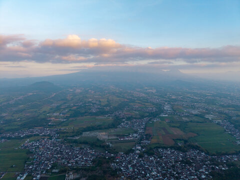 Aerial view of Malang city nestled among lush green fields and agricultural lands with a distant mountain peak under a soft sky in Malang, East Java, Indonesia.