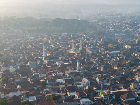 Aerial view of the dense urban landscape featuring prominent mosques with green domes and minarets amidst residential rooftops in the morning light in Malang, East Java, Indonesia.