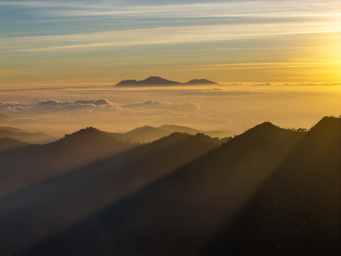 Aerial view of Cemorolawang during sunset with dramatic light rays illuminating the mountain peaks, rolling hills, and a sea of clouds in Kecamatan Sukapura, Jawa Timur, Indonesia.