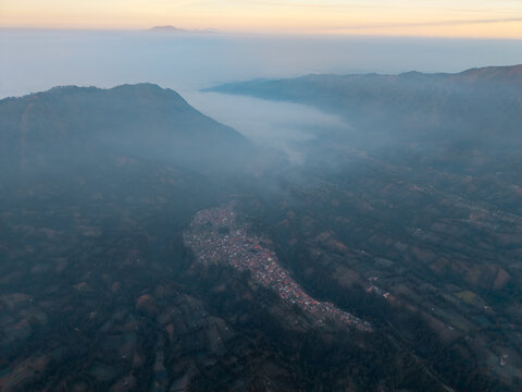 Aerial view of Wonotoro village nestled in a valley surrounded by misty mountains during sunset in Sukapura, East Java, Indonesia.