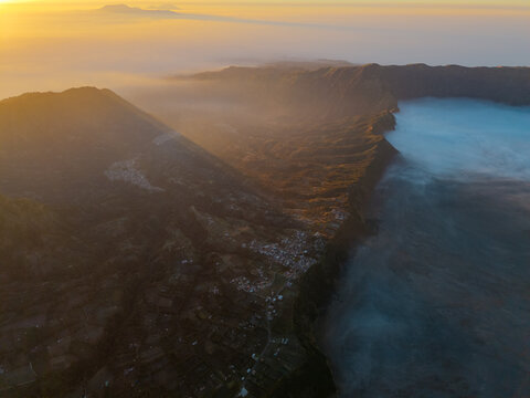 Aerial view of Mount Bromo at sunrise with golden light illuminating the volcanic crater and sea of clouds over the rugged landscape in Tosari, East Java, Indonesia.