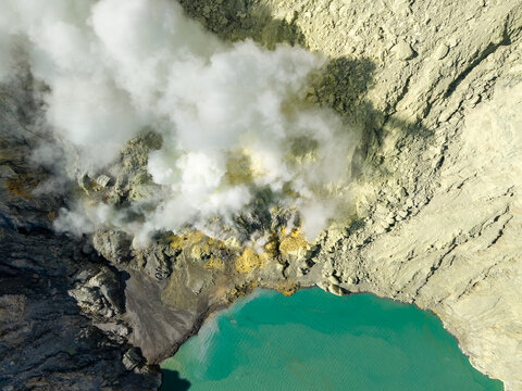Aerial view of Mount Ijen sulfur mining operations with thick volcanic smoke rising from the crater near the turquoise acidic lake in Ijen, East Java, Indonesia.
