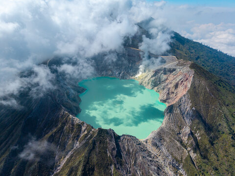 Aerial view of Ijen crater lake with its turquoise water surrounded by rugged volcanic slopes and clouds in Ijen, East Java, Indonesia.