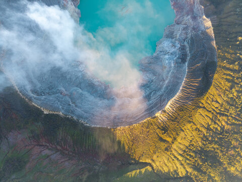 Aerial view of Ijen crater lake with vibrant turquoise water, surrounded by volcanic crater rims, rising steam, and rugged terrain in golden hour light in Ijen, East Java, Indonesia.