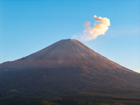 Aerial view of Mount Semeru peak with a volcanic smoke plume rising into a clear blue sky above lush forested slopes in Kecamatan Pronojiwo, Jawa Timur, Indonesia.