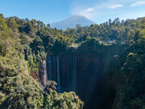 Aerial view of Tumpak Sewu Waterfall cascading into a lush canyon with Mount Semeru volcano in the background in Kecamatan Pronojiwo, Jawa Timur, Indonesia.