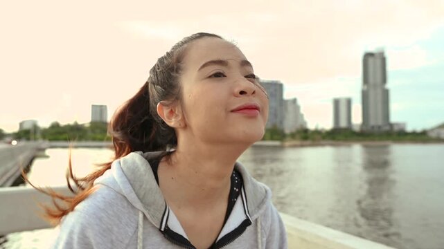 A woman in sportswear stands by the waterfront wearing a gray zip hoodie and athletic leggings. The woman faces the camera near a metal railing, with water and distant city buildings behind her.