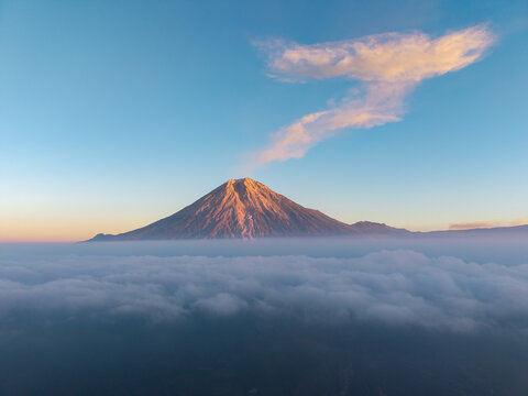 Aerial view of Semeru volcano rising above a thick layer of clouds during golden hour with soft light on the peak in Pasrujambe, East Java, Indonesia.